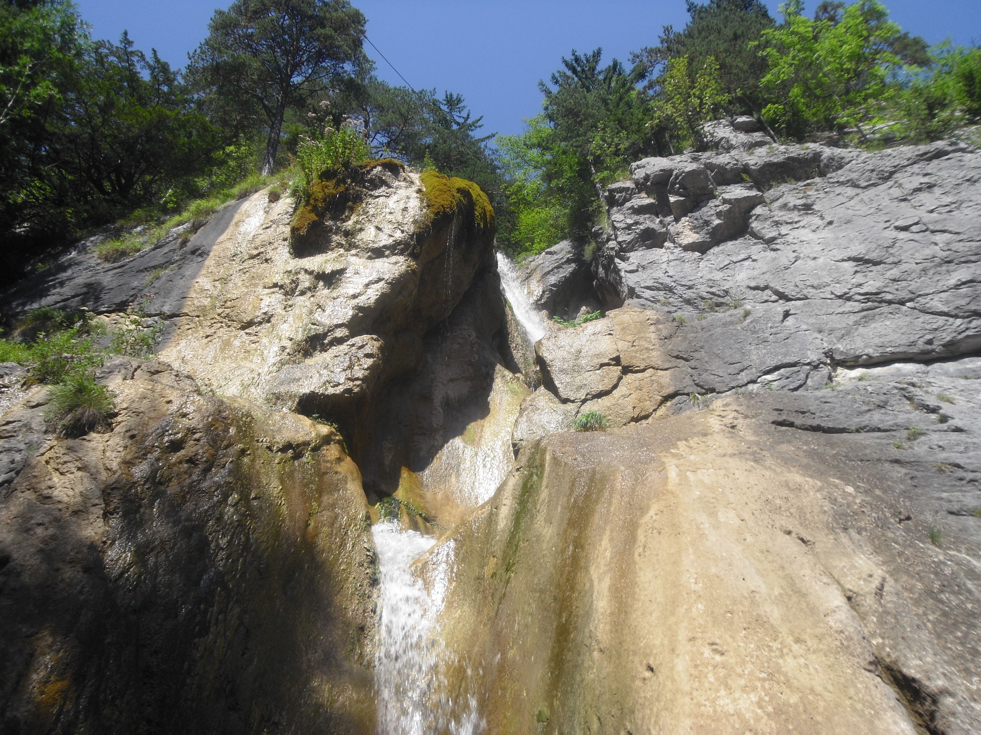 Wasserfall in felsiger Landschaft mit Bäumen und blauem Himmel.