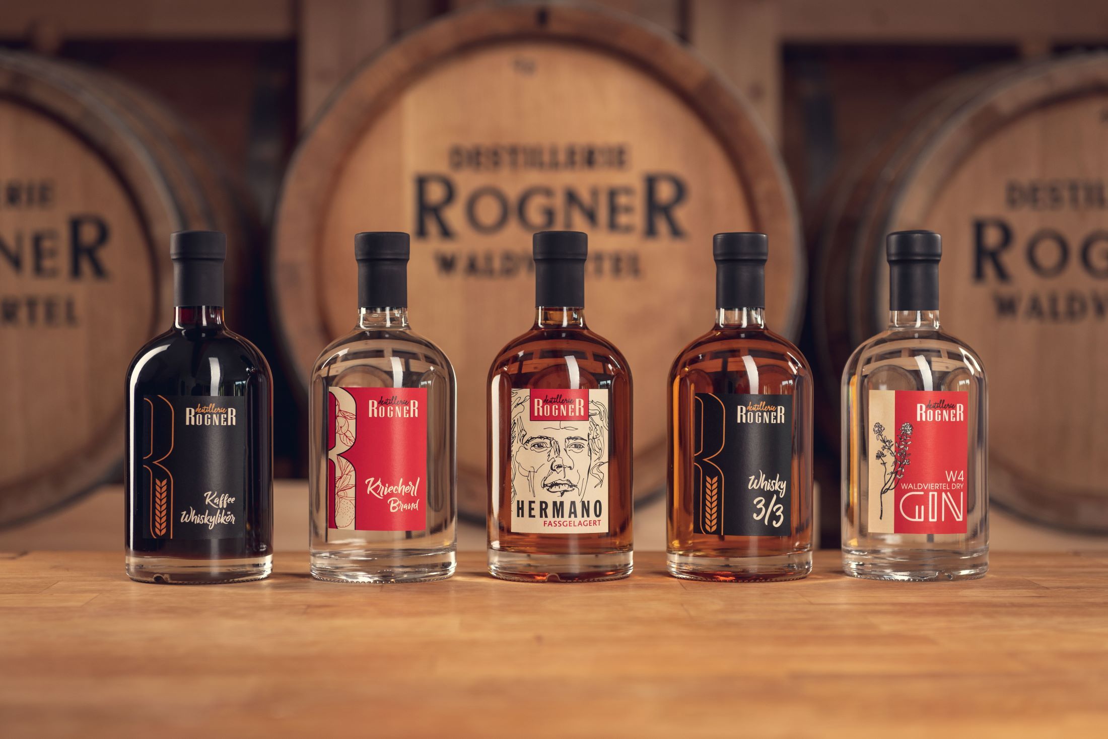 Fünf Flaschen Spirituosen der Destillerie Rogner vor Holzfässern.