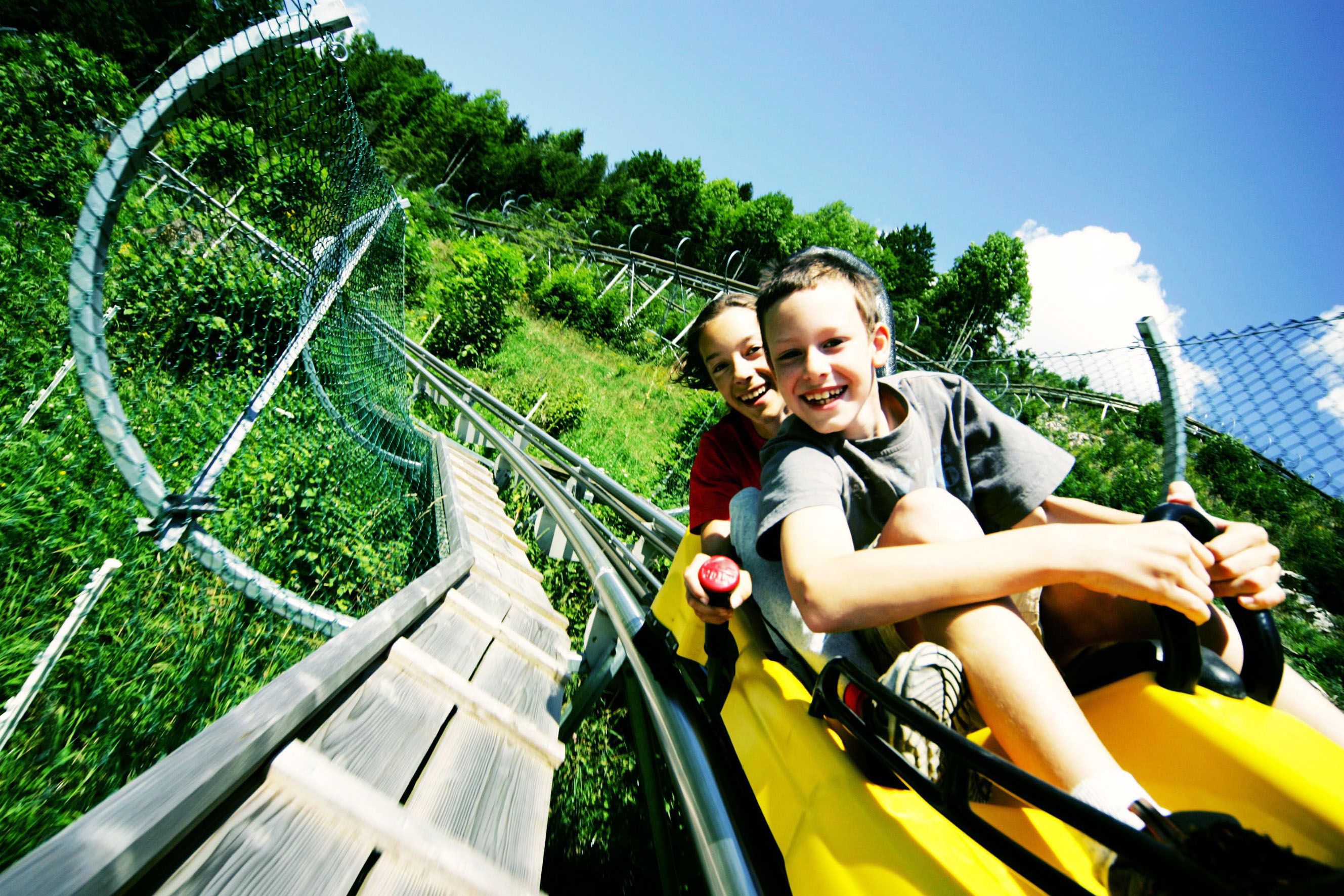 Zwei Kinder fahren lachend auf einer Sommerrodelbahn durch grüne Landschaft.