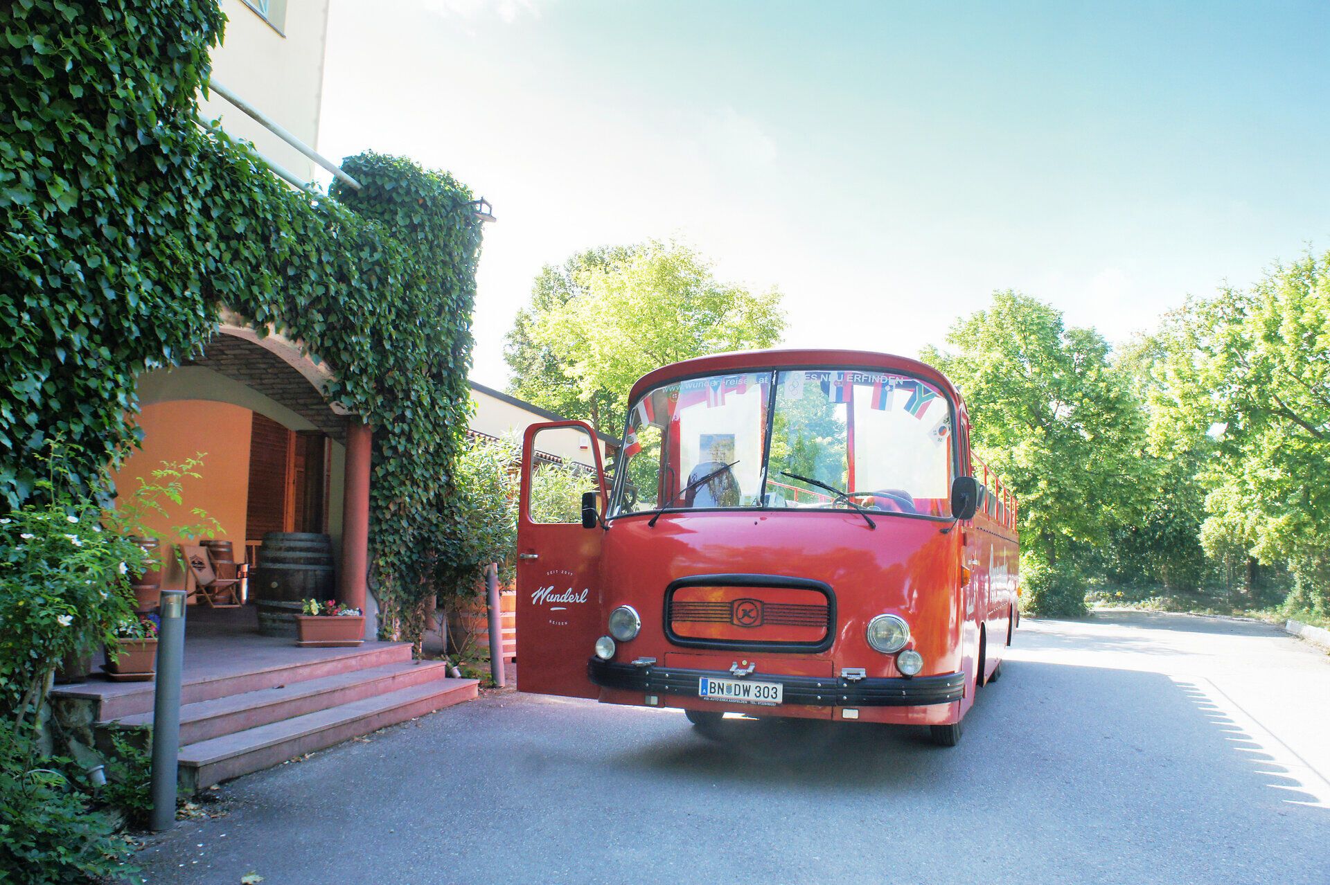 Roter Oldtimerbus steht vor Heurigen-Weingut mit geöffnete Türe an sonnigem Tag und wartet auf den Einstieg der Gäste.
