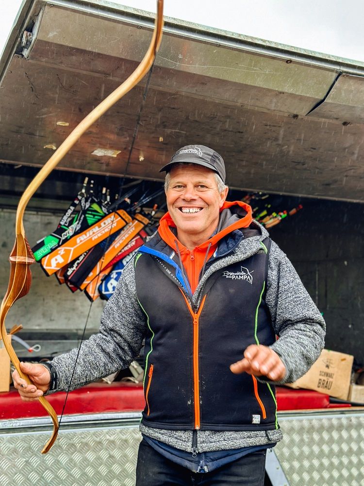 Ein lächelnder Mann hält einen Bogen vor einem Anhänger mit Pfeilen.