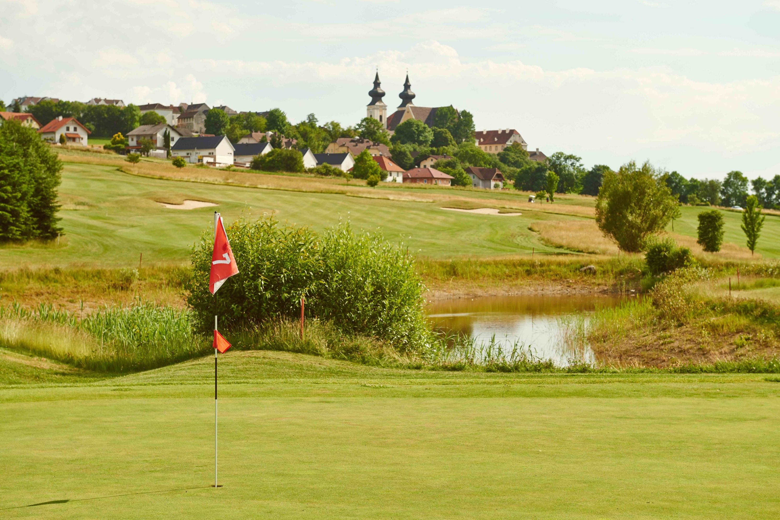 Golfplatz mit Fahne im Vordergrund, Dorf und Kirche im Hintergrund.