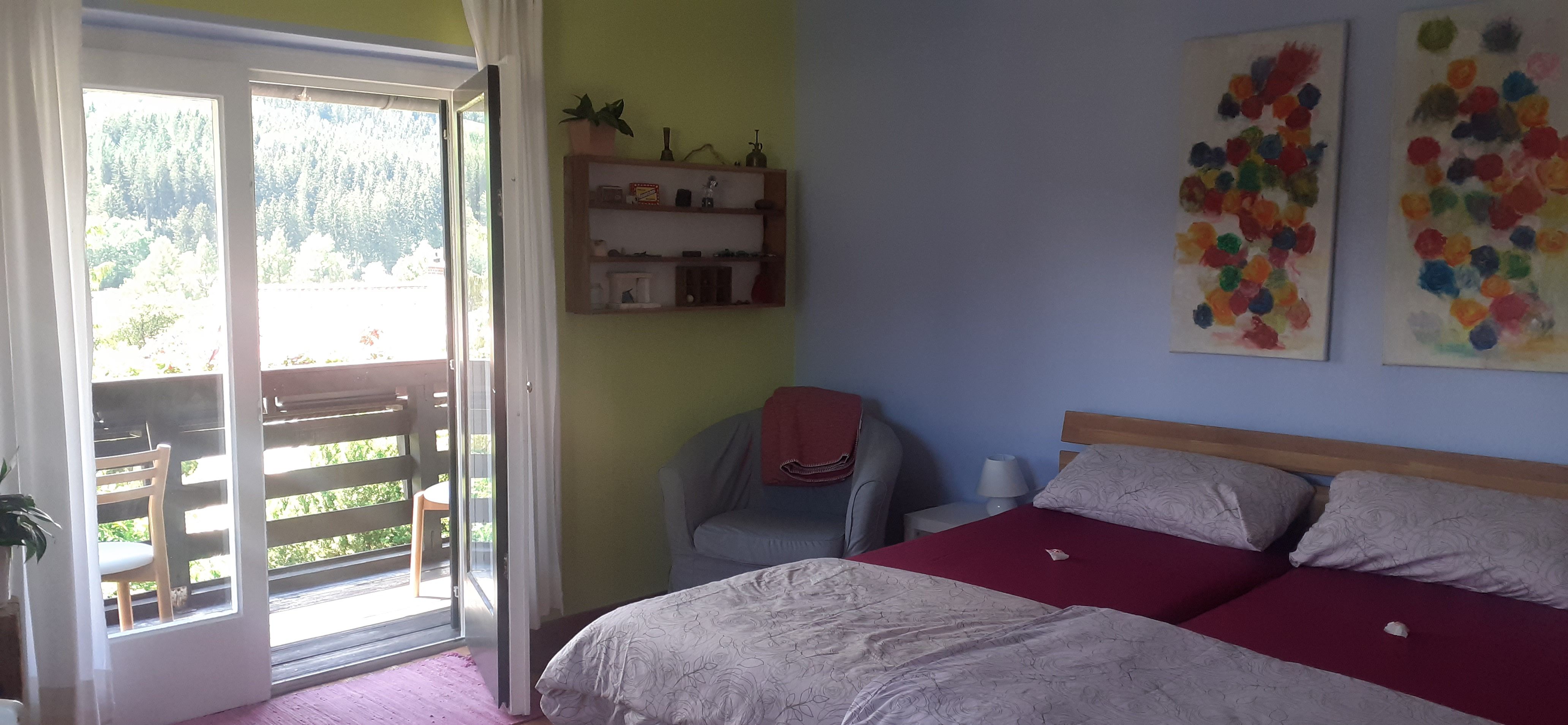 Ein gemütliches Doppelzimmer mit zwei Betten, einem Balkon mit Blick auf Bäume und bunte Kunstwerke an der Wand.