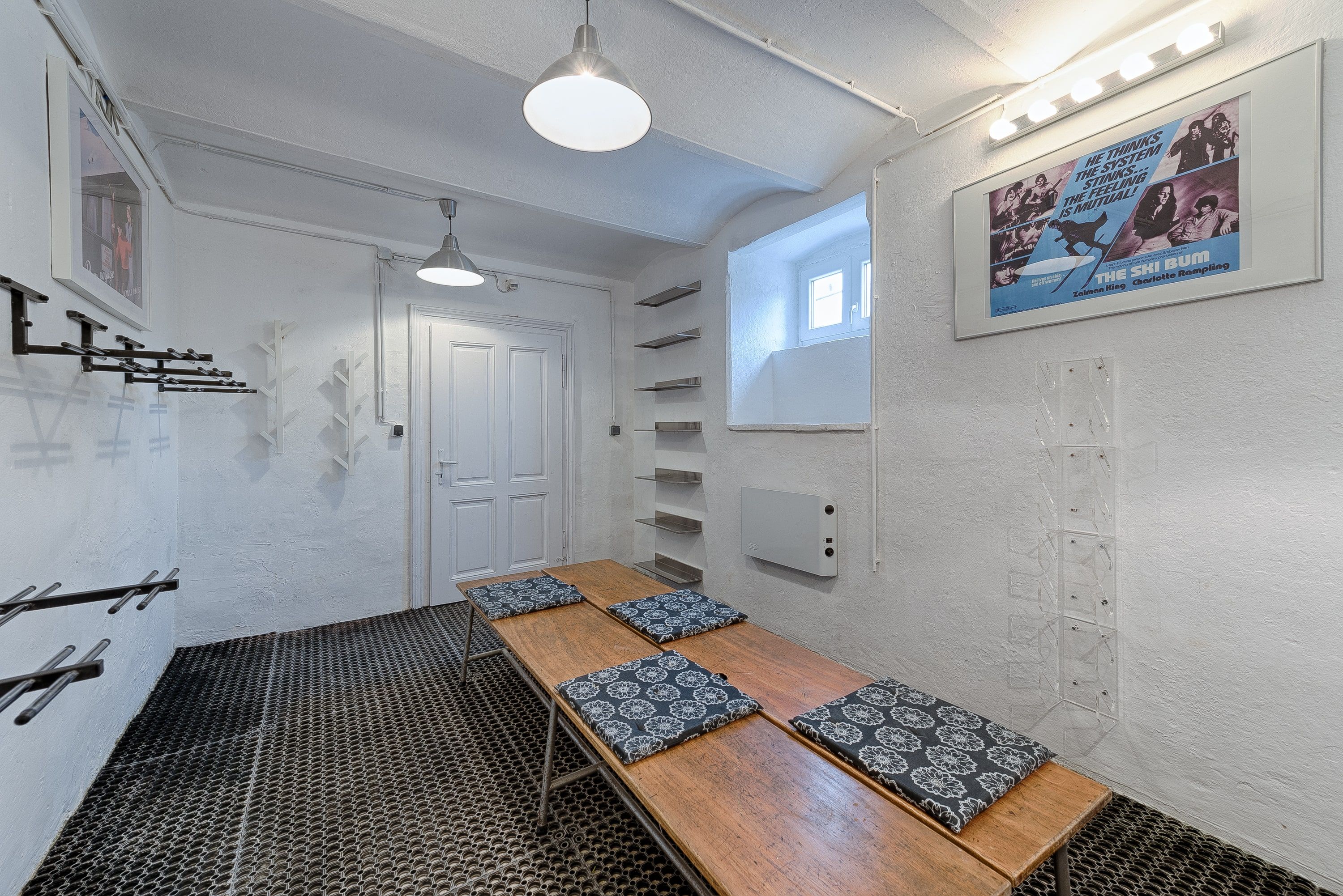 Ein Skikeller mit weißen Wänden, Skihalterungen, einer Holzbank mit Kissen und einem Filmplakat an der Wand.