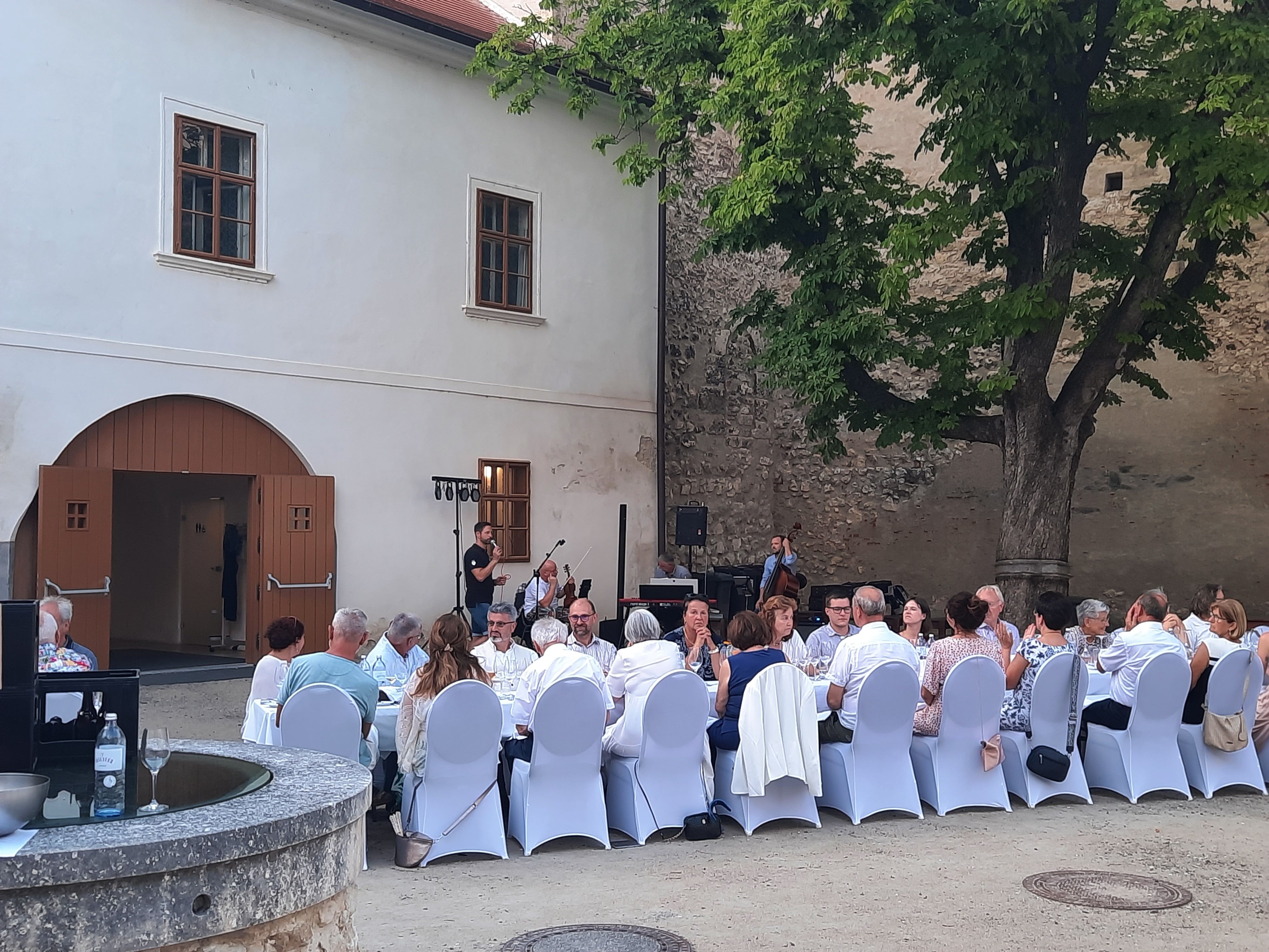 Menschen sitzen an einem langen Tisch im Freien, umgeben von alten Gebäuden und einem Baum.