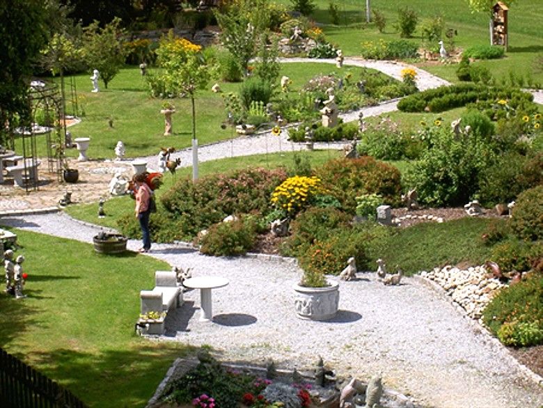 Ein gepflegter Schaugarten mit geschwungenen Wegen, Blumenbeeten und Gartenfiguren. Eine Person betrachtet die Pflanzen.