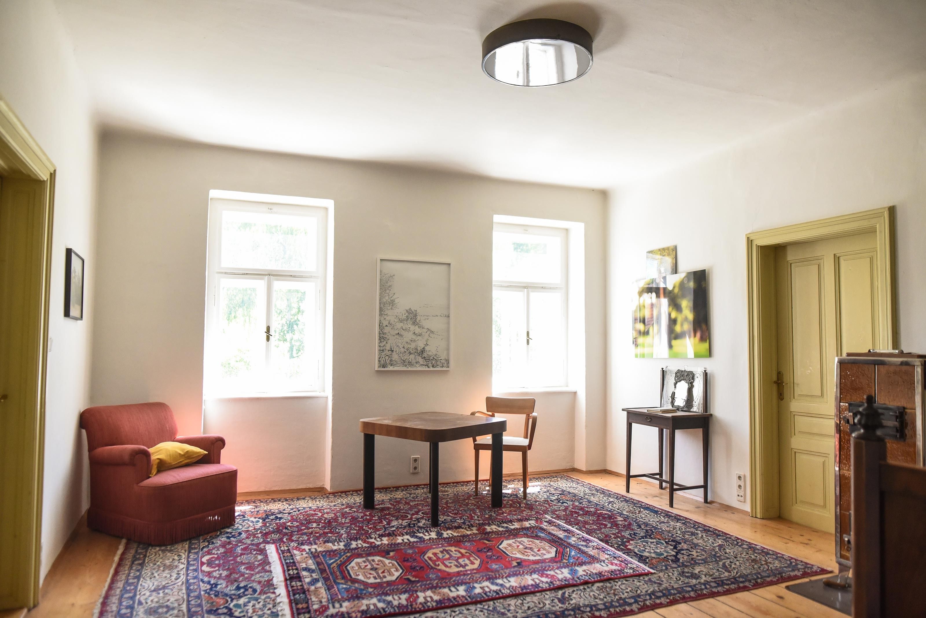 Ein helles Wohnzimmer mit einem roten Sessel, einem Holztisch und einem Stuhl auf einem orientalischen Teppich.