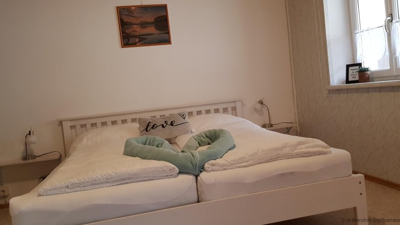 Zimmer, © mittendrin Dorfpension zum Ehn Weissesdoppelbett mit einer Decke, welche ein Herz formt. Über dem Bett hängt auf einer weißen Wand ein rotes Bild.