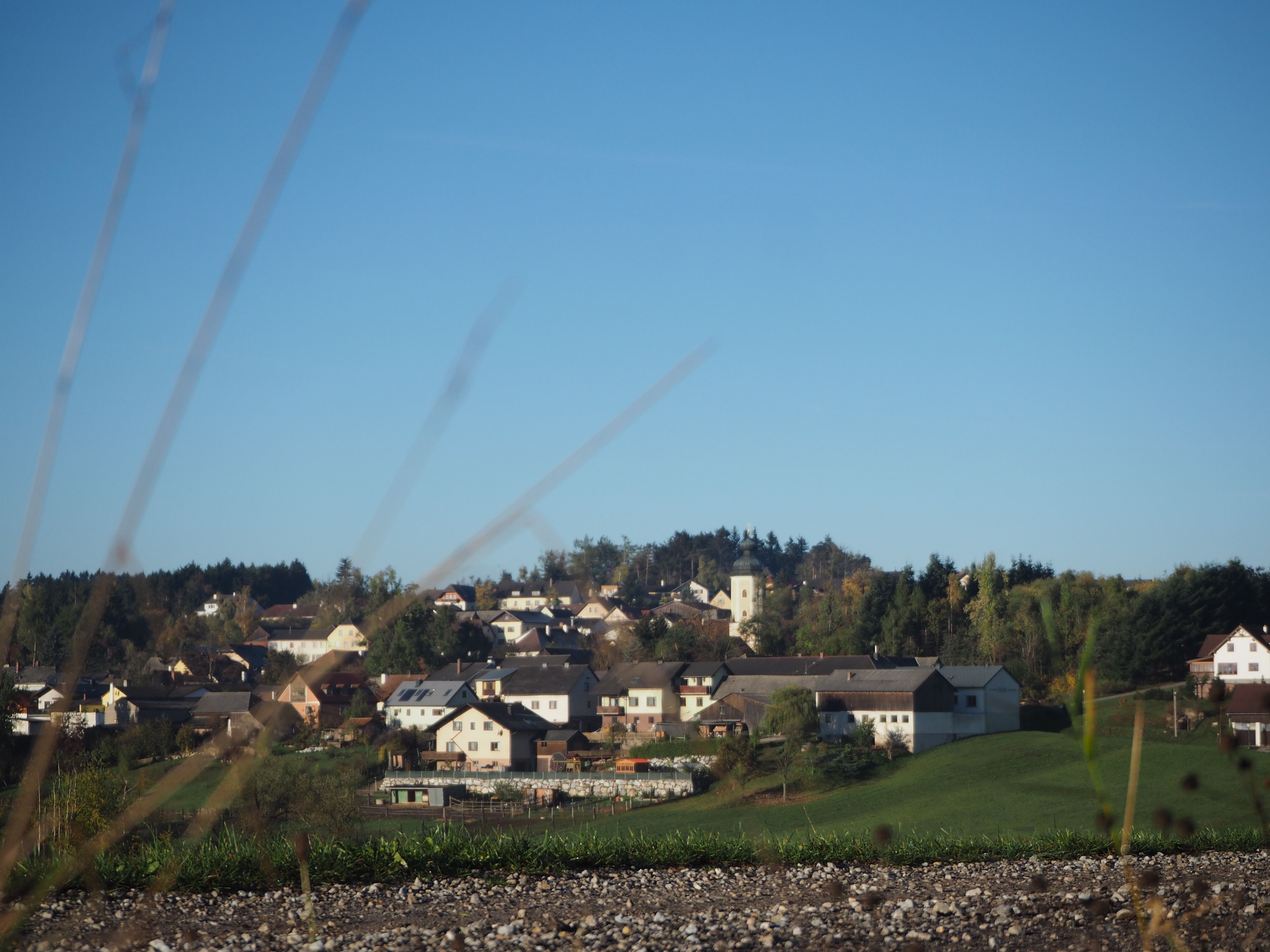 Ländliche Siedlung mit Kirche und Häusern auf einem Hügel, umgeben von Bäumen und Wiesen unter klarem Himmel.