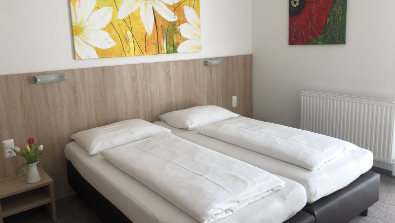 Ein schlichtes Hotelzimmer mit zwei Einzelbetten, einem Nachttisch mit Blumen und zwei bunten Gemälden an der Wand.