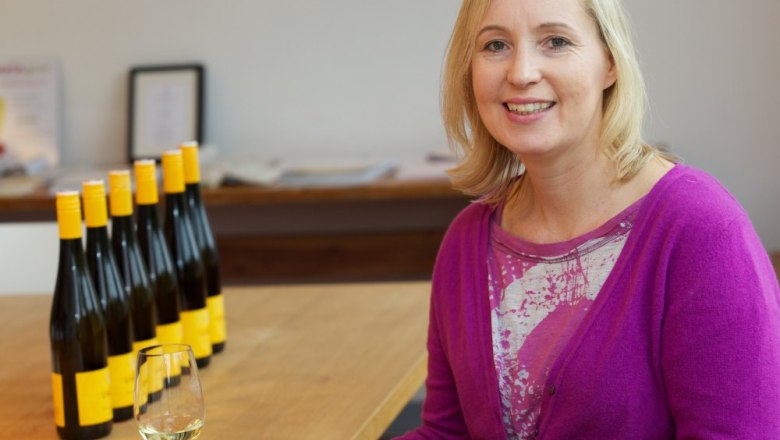 Frau in lila Pullover mit Weinglas, Flaschen im Hintergrund.