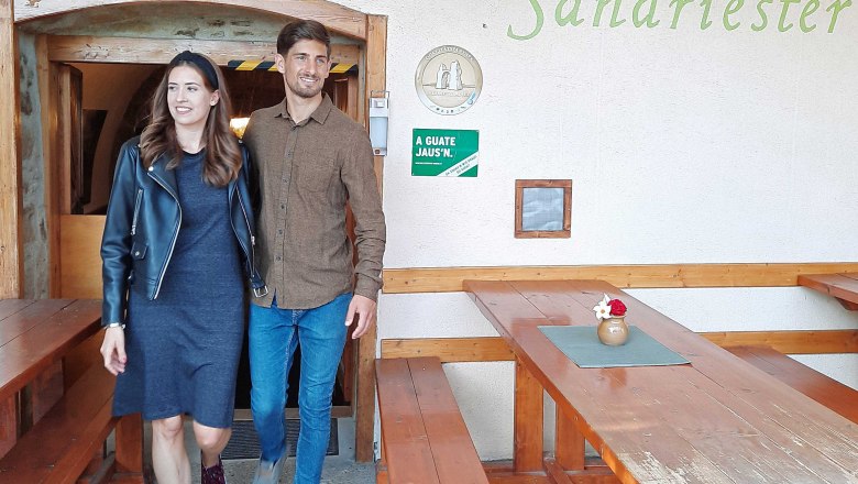 Ein Mann und eine Frau verlassen eine Buschenschank mit dem Namen Gratzer-Sandriester. Sie l&auml;cheln und gehen an Holzb&auml;nken vorbei.