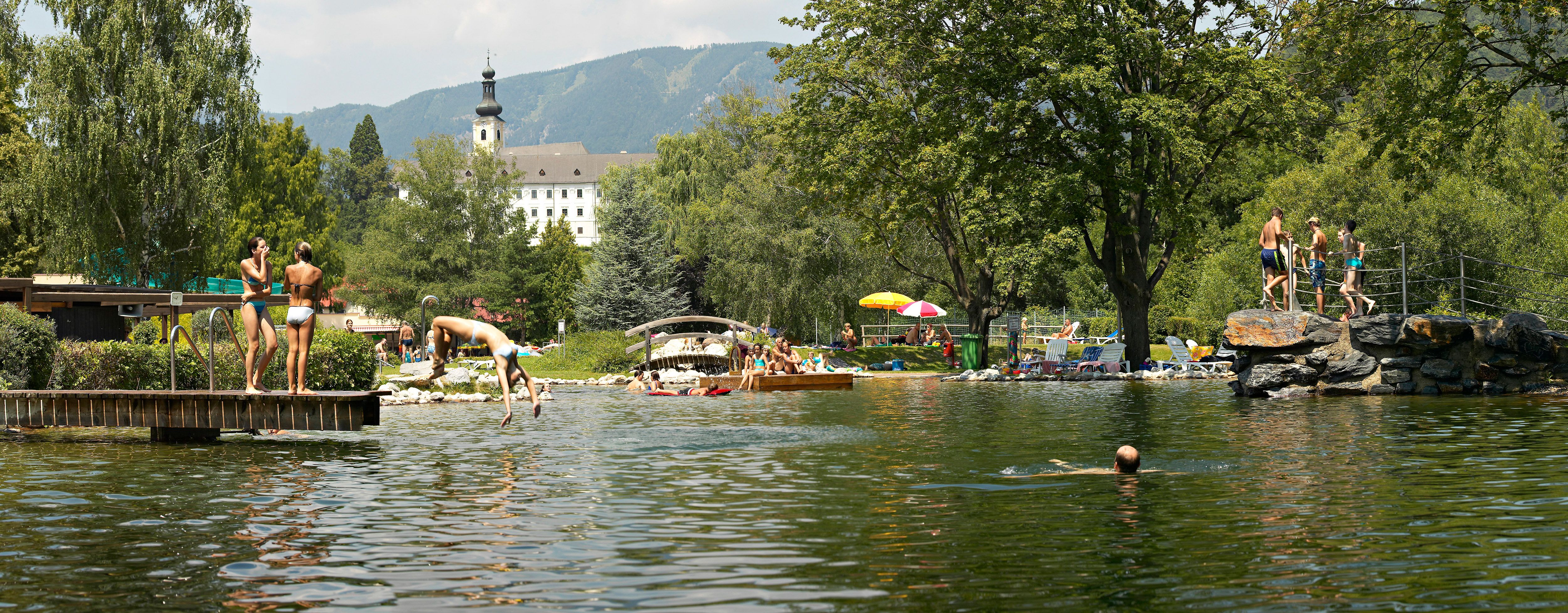 Menschen genießen einen sonnigen Tag im Naturbad Gloggnitz mit Schwimmen und Sonnenbaden, umgeben von Bäumen und Bergen.