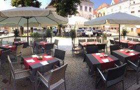 Außenterrasse eines Cafés mit Tischen und Sonnenschirmen auf einem gepflasterten Platz.