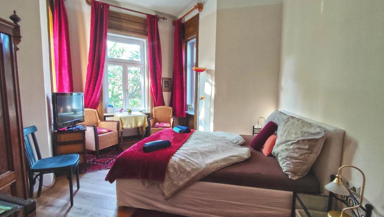 Gemütliches Schlafzimmer mit Doppelbett, roten Vorhängen und Sesseln vor einem Fenster.