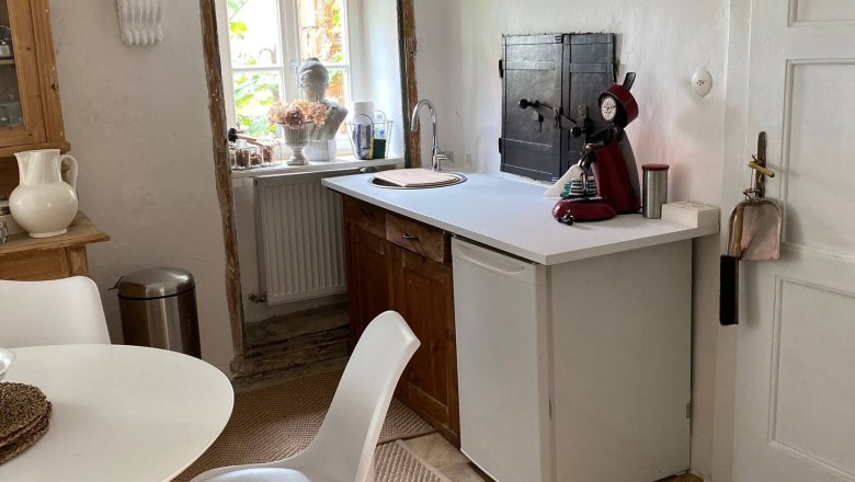 Küchenbereich mit kleinem Tisch, Stuhl, Spüle und Kaffeemaschine. Fenster mit Dekoration und helle Wände.