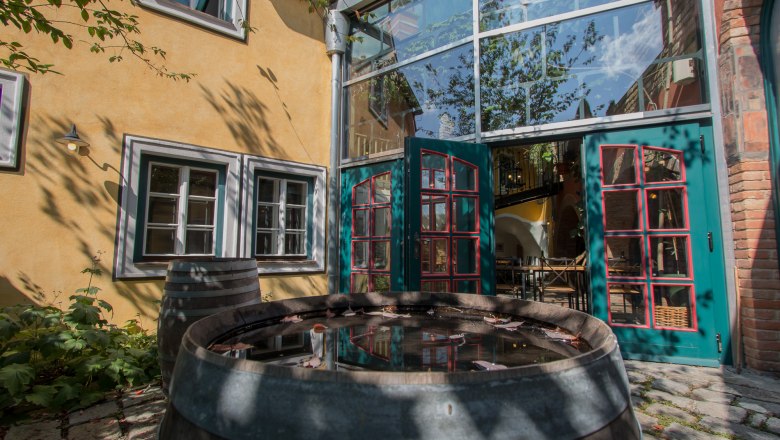 Ein Innenhof mit gelber Fassade, grünen Türen und einem Holzfass mit Wasser. Schatten von Bäumen fallen auf die Wand.