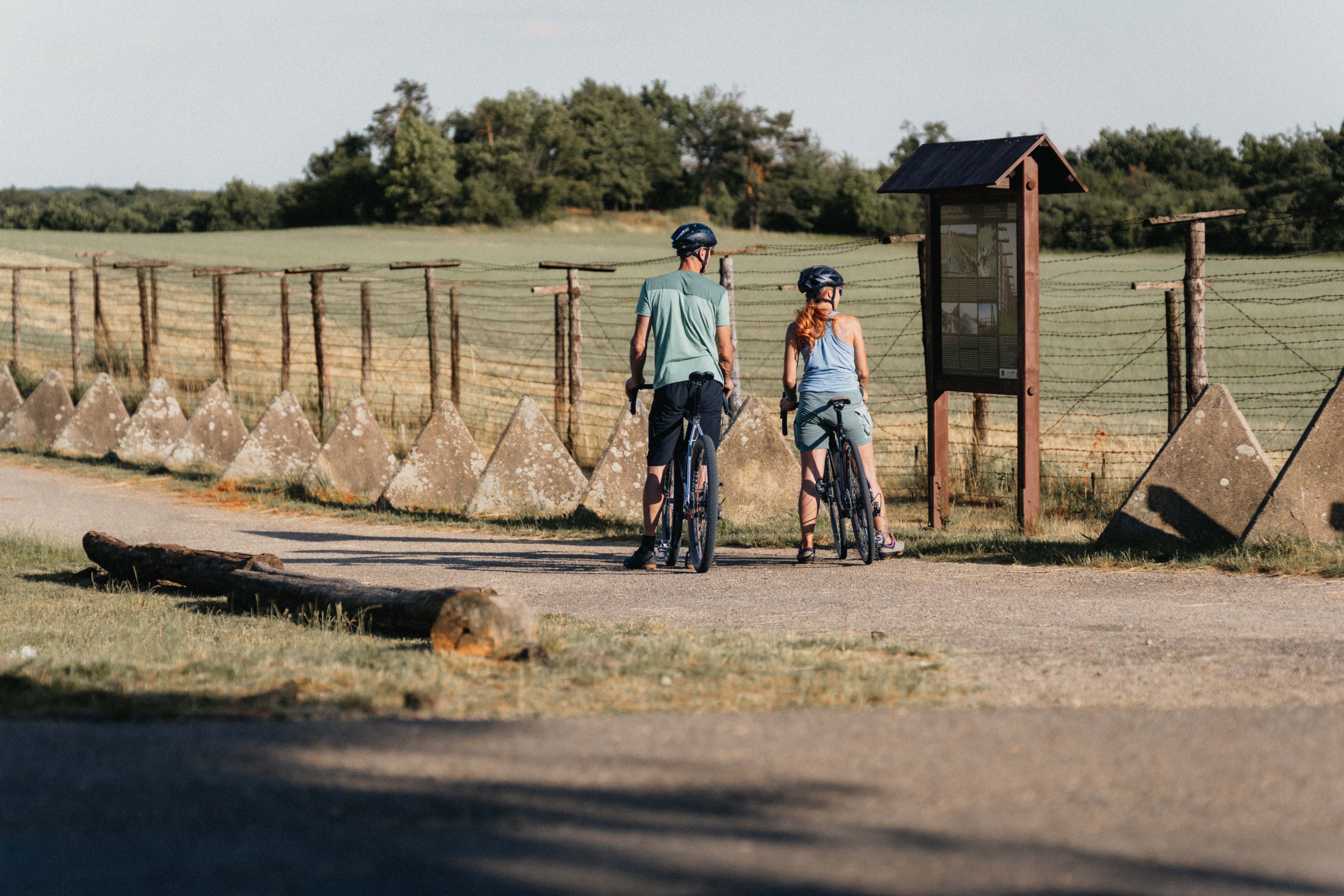 Zwei Radfahrer stehen vor einem historischen Grenzzaun mit Informationstafel.