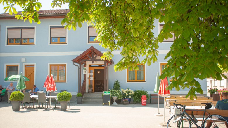 Ein blaues Gasthaus mit Sonnenschirmen und Tischen im Freien, umgeben von grünen Bäumen.