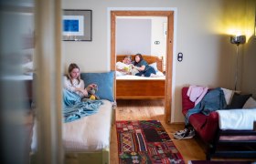 Innenansicht einer Ferienwohnung mit zwei Zimmern. Im Vordergrund sitzt ein Mädchen auf einem Bett, im Hintergrund lesen zwei Personen auf einem anderen Bett.