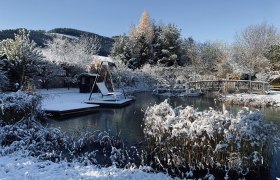 Winterlicher Garten mit Teich, Br&uuml;cke und Schaukel im Schnee.
