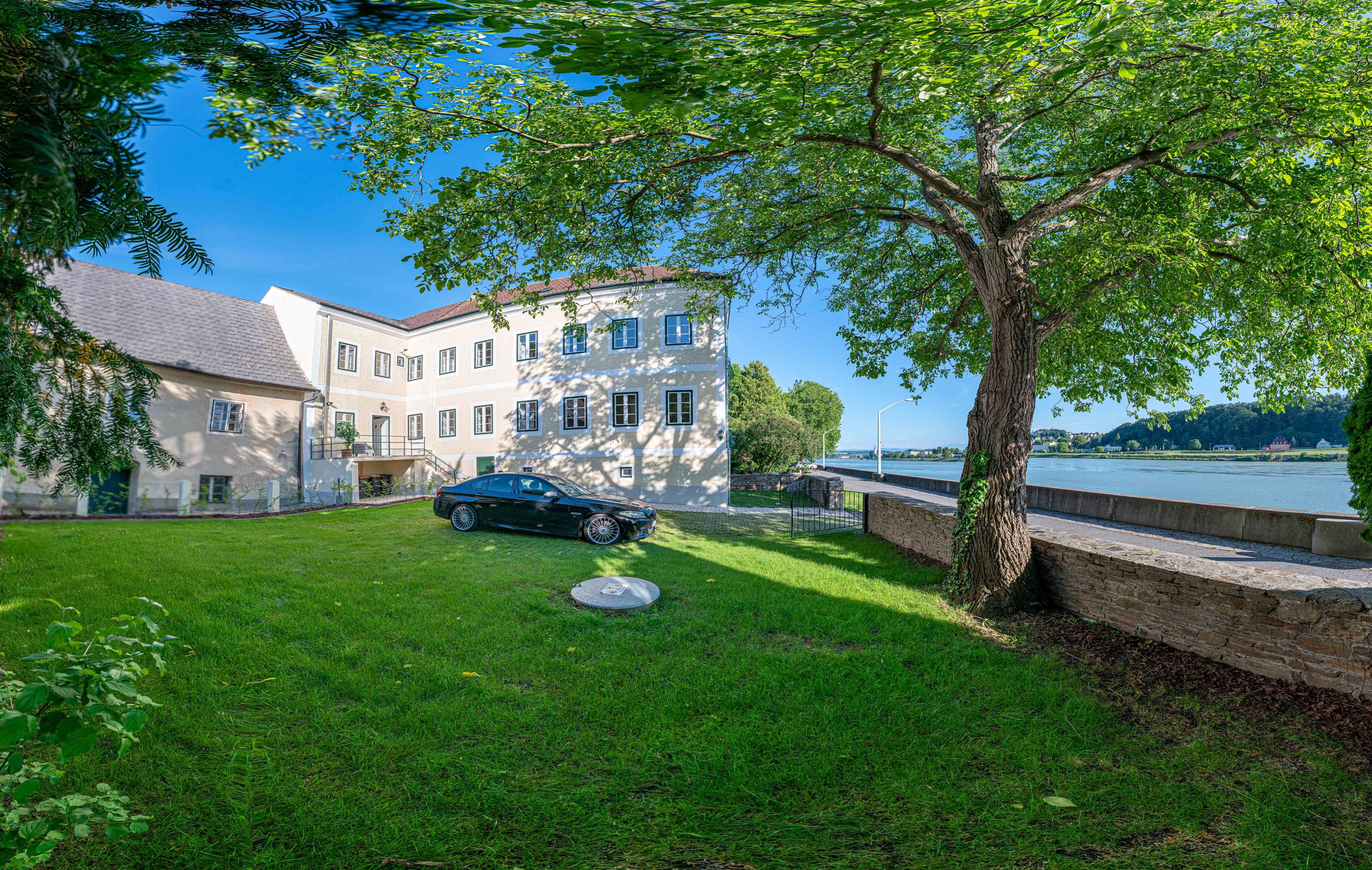 Außenansicht eines alten Postamts mit Baum und Auto im Vordergrund, Donau im Hintergrund.