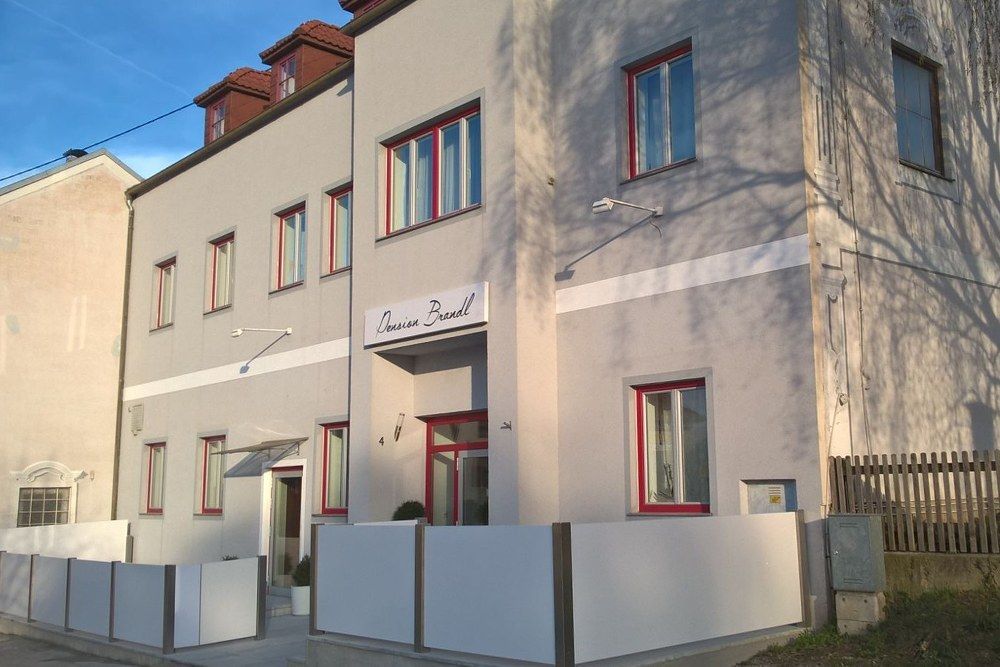 Außenansicht der Pension Brandl mit grauer Fassade und roten Fensterrahmen.