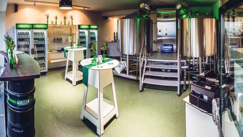 Innenansicht einer kleinen Brauerei mit Edelstahlkesseln und Kühlschränken voller Getränke.