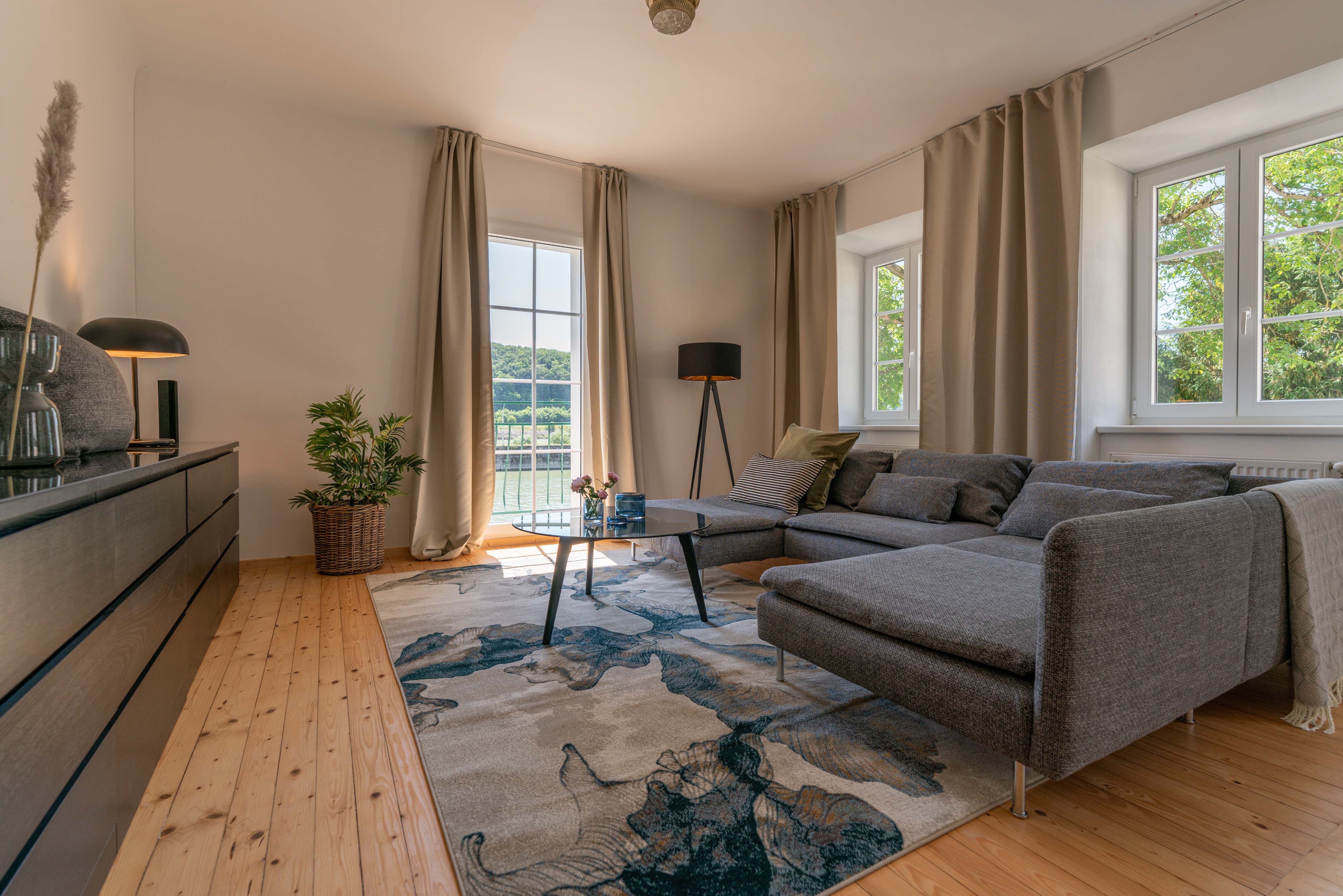 Modernes Wohnzimmer mit grauem Sofa, Holzboden und großem Fenster mit Blick ins Grüne.
