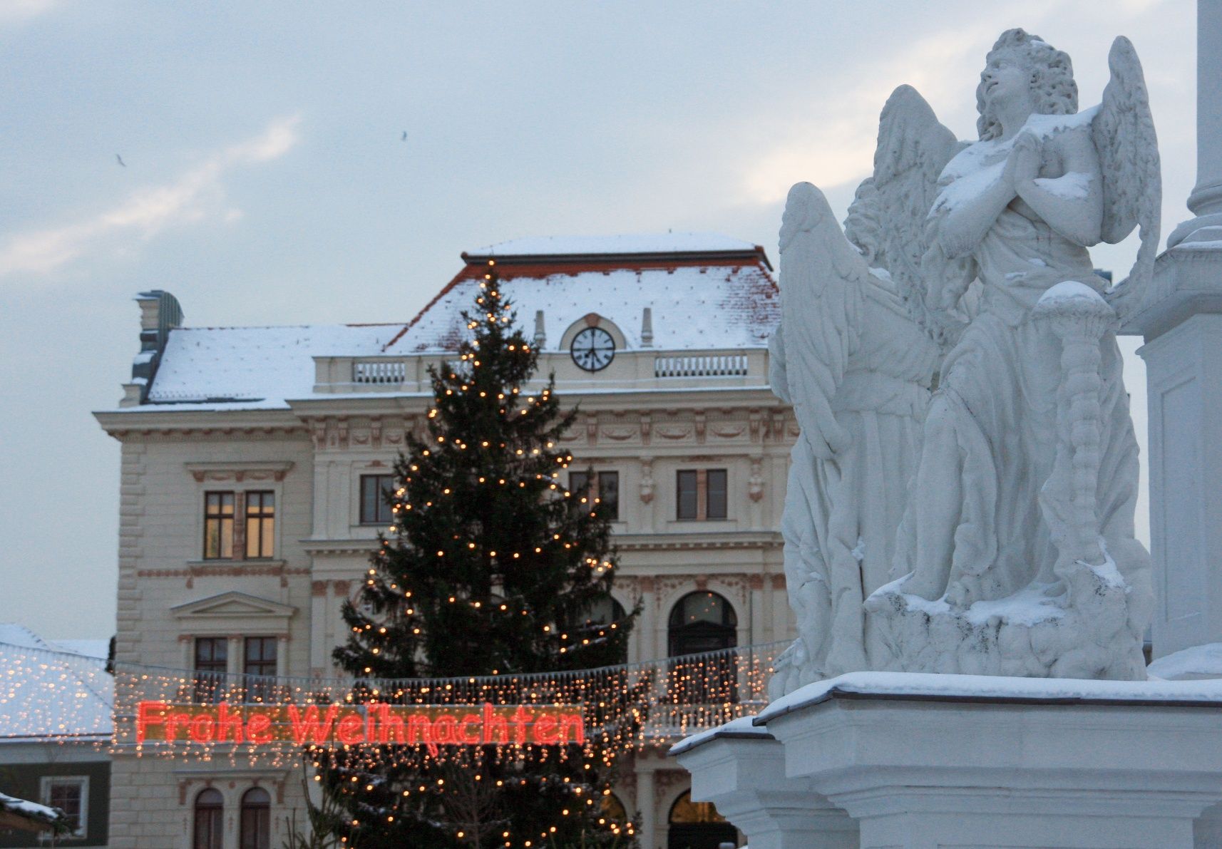 Weihnachtlich geschmückter Platz mit Engelstatue, Weihnachtsbaum und Lichterkette mit der Aufschrift 'Frohe Weihnachten'.