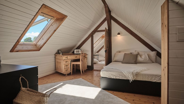 Gem&uuml;tliches Dachzimmer mit Holzbalken, Bett, Schreibtisch und Dachfenster.