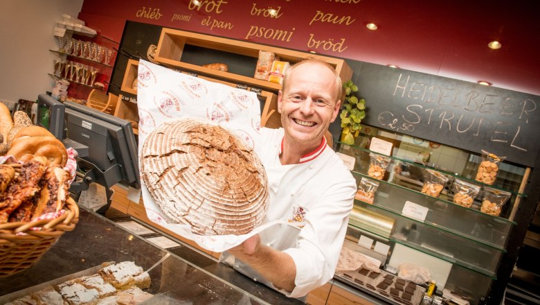 Ein B&auml;cker in einer B&auml;ckerei h&auml;lt ein gro&szlig;es, rundes Brot l&auml;chelnd in die Kamera. Im Hintergrund sind Regale mit Backwaren und eine Tafel mit der Aufschrift 'Heidelbeer Strudel'.