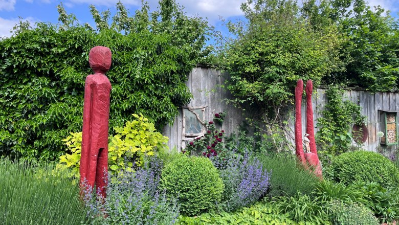 Garten mit roten Skulpturen, umgeben von gr&uuml;nen Pflanzen und einer Holzwand im Hintergrund.