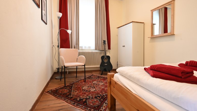 Kleines Zimmer mit Bett, Teppich, Stuhl, Stehlampe, Gitarre und Schrank.