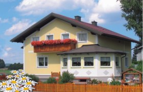 Ein gelbes Haus mit rotem Dach und Blumenkasten auf dem Balkon, umgeben von einem Holzzaun und Gänseblümchen im Vordergrund.