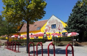 Ein gelbes Geb&auml;ude mit Wandmalerei, umgeben von B&auml;umen und roten Sonnenschirmen.