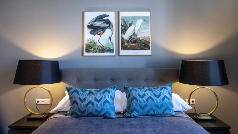 Doppelzimmer mit grauem Bett, blauen Kissen, zwei Nachttischlampen und Vogelbildern an der Wand.