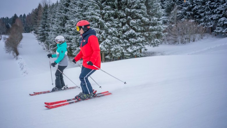 Zwei Skifahrer auf einer verschneiten Piste, umgeben von schneebedeckten B&auml;umen.