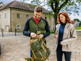 Ein Mann und eine Frau stehen drau&szlig;en vor dem Schloss Katzelsdorf. Der Mann packt einen gr&uuml;nen Rucksack, w&auml;hrend die Frau zuschaut. Beide tragen Outdoor-Kleidung.