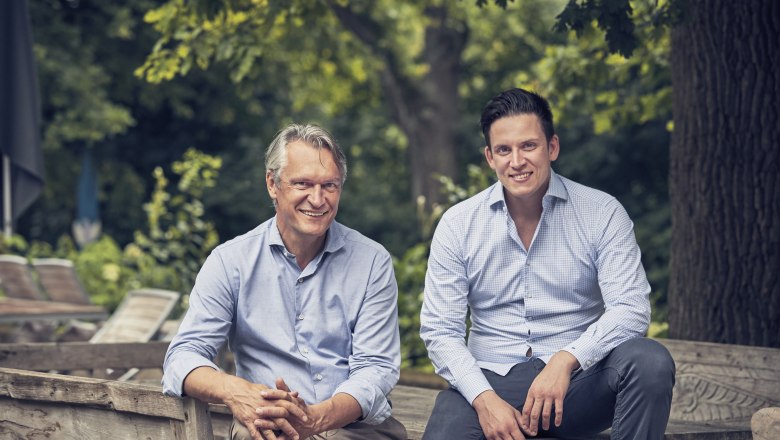 Zwei Männer sitzen auf einer Holzbank im Freien, umgeben von Bäumen.