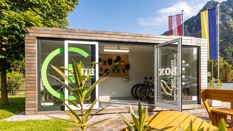Eine moderne Fahrradbox mit geöffneten Türen, Fahrrädern und Helmen, umgeben von Natur und Flaggen.