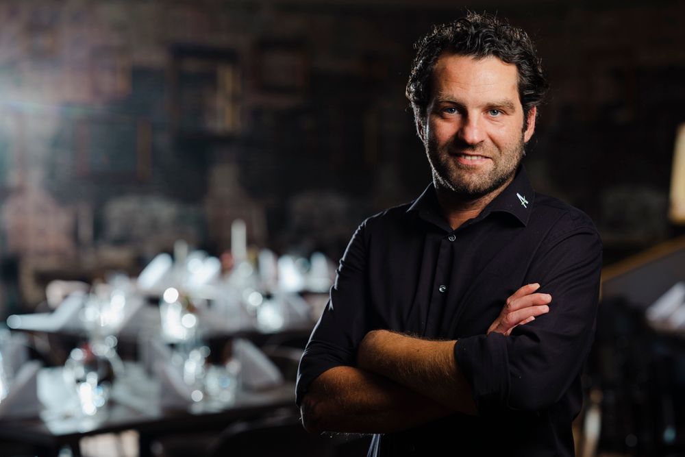 Mann in schwarzem Hemd steht vor elegant gedecktem Tisch in dunklem Raum.