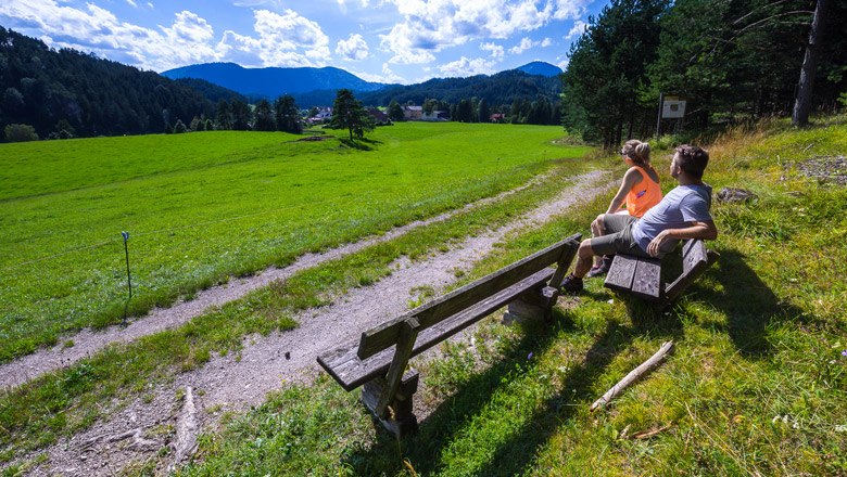 Zwei Personen sitzen auf einer Bank und blicken auf eine gr&uuml;ne Wiese mit Bergen im Hintergrund.