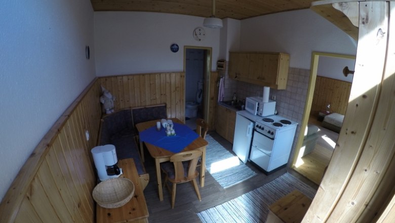 Wohnk&uuml;che in einer Ferienwohnung mit Holzm&ouml;beln, Esstisch, K&uuml;chenzeile und Zugang zu einem Schlafzimmer.