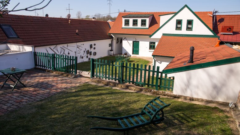Ein traditioneller Winzerhof mit roten Ziegeld&auml;chern, gr&uuml;nen Z&auml;unen und Gartenm&ouml;beln auf einer Wiese.