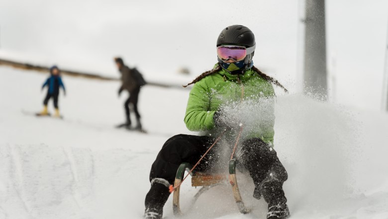 Person mit grünem Anorak und Helm fährt auf einem Schlitten im Schnee.