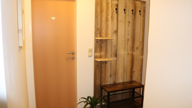 Eingangsbereich mit Holzt&uuml;r, Garderobe aus Holz mit Haken, Bank und Pflanze.