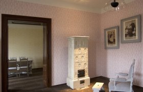 Innenansicht eines Raumes mit antikem Ofen, zwei gerahmten Bildern und einem Stuhl.