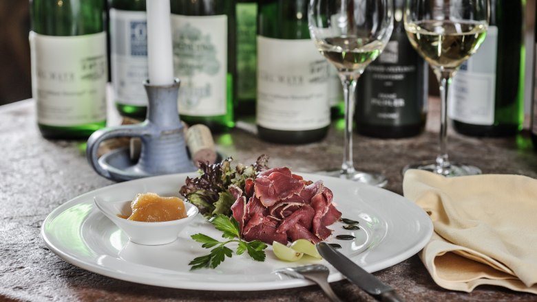 Ein Teller mit Schinken, Salat und Chutney, umgeben von Weinflaschen und Gl&auml;sern auf einem Tisch.