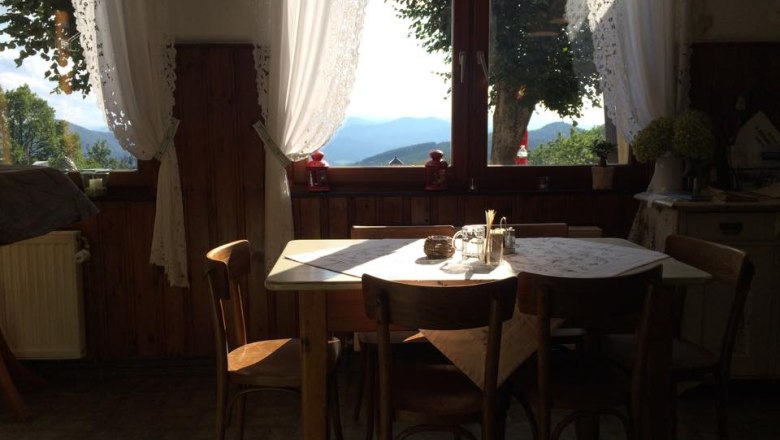 Ein gem&uuml;tlicher Essbereich mit Holztisch und St&uuml;hlen vor einem gro&szlig;en Fenster mit Blick auf Berge und B&auml;ume.
